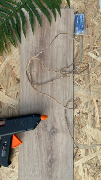 Heißklebepistole, Schnur und Batterien auf Laminatboden mit Farnblatt daneben.