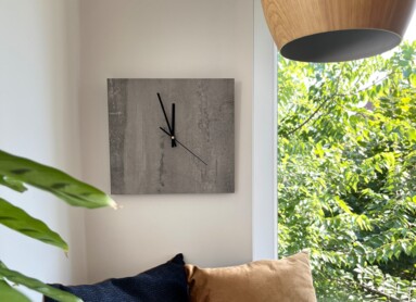 Gemütliche Ecke mit Kissen, Wanduhr und Pflanzen, neben einem Fenster mit Blick ins Grüne.