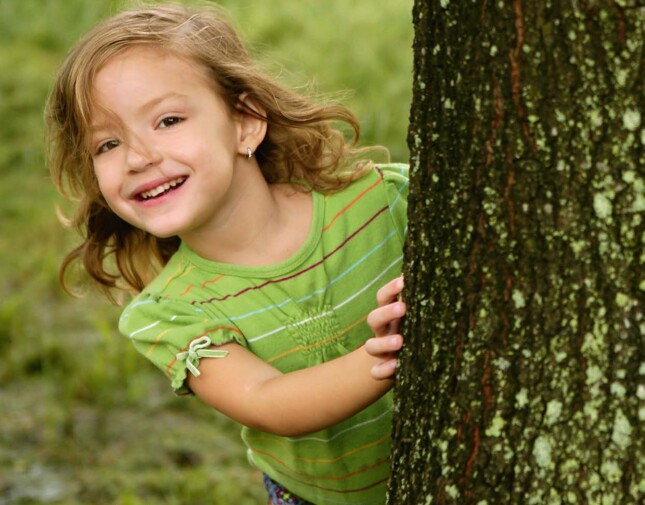 A child smiling behind a tree in a grassy area, wearing a green striped shirt and jeans.