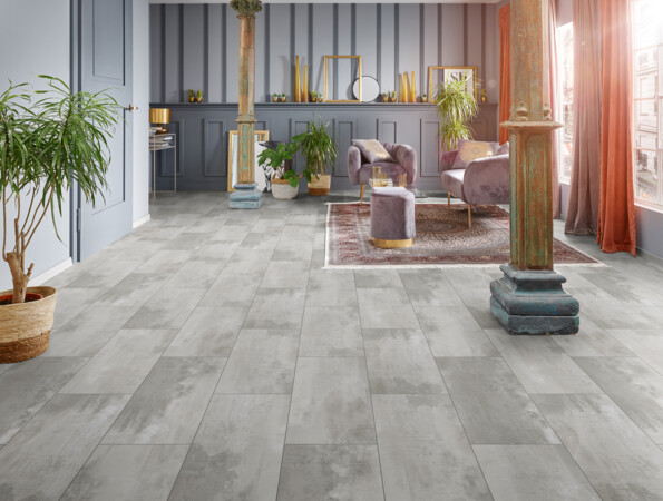 Spacious living room with grey laminate flooring, vintage columns, and plush seating, accented by indoor plants and warm sunlight.