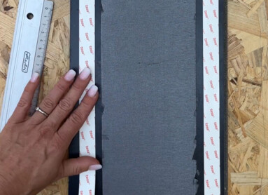 Eine Person bringt doppelseitiges Klebeband auf einer Laminatplatte an, neben einem Lineal auf einem Holzuntergrund.