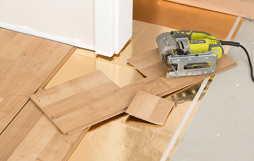 Eine Stichsäge liegt auf Laminatdielen, die auf einer goldenen Unterlage verlegt werden, und zeigt einen Bodenbelagsprozess.