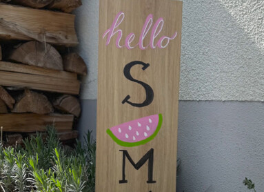 Holzschild im Garten mit der Aufschrift "hello SUMMER" und einer Wassermelonengrafik. Im Hintergrund sind Pflanzen und Holzstapel sichtbar.