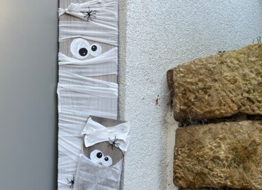 Eine DIY-Halloween-Dekoration mit Mumienfiguren aus Holz und Bändern, dekoriert mit Spinnen und Augen, neben einer Laterne im Freien.