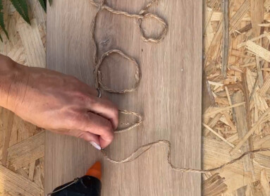 Hand gestaltet ein Seilbild auf Laminatboden mit Heißklebepistole, neben Farnblättern.