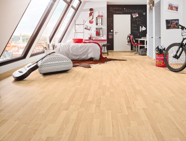 Attic bedroom with light laminate flooring, featuring a bed, desk, guitar, and bicycle near large windows.