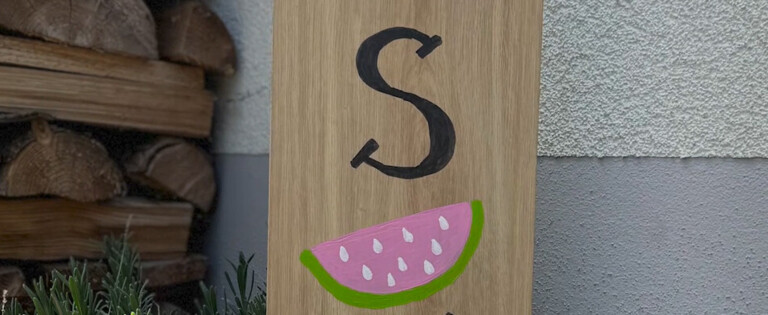 Holzschild im Garten mit der Aufschrift "hello SUMMER" und einer Wassermelonengrafik. Im Hintergrund sind Pflanzen und Holzstapel sichtbar.