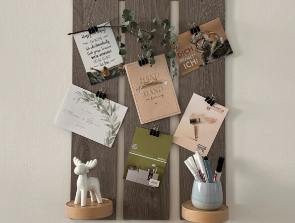 Wanddekoration mit Fotos und Karten auf Holzpaneelen, darunter ein Regal mit Keramikfigur und Stiftehalter.