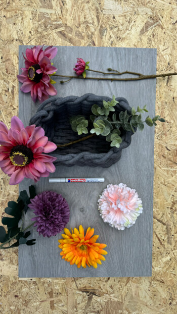 Verschiedene Kunstblumen und ein graues Strickkörbchen auf einem Laminatbodenmuster.