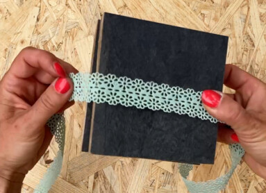 Eine Person hält ein schwarzes Buch mit dekorativem Band auf einem Holzuntergrund.