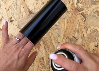 Eine Hand sprüht Farbe auf ein schwarzes Rohr, das auf einem Holzuntergrund liegt.