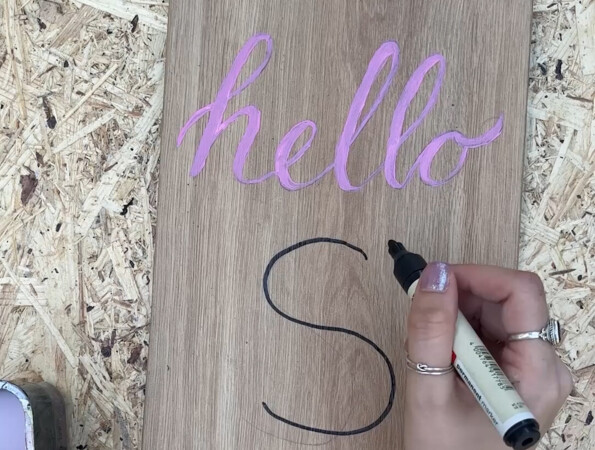 Eine Hand schreibt "S" auf ein Holzbrett mit "hello" in rosa Schrift.