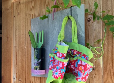Bunte Gartentasche und Werkzeuge hängen an einer Holztür, umgeben von Blättern.