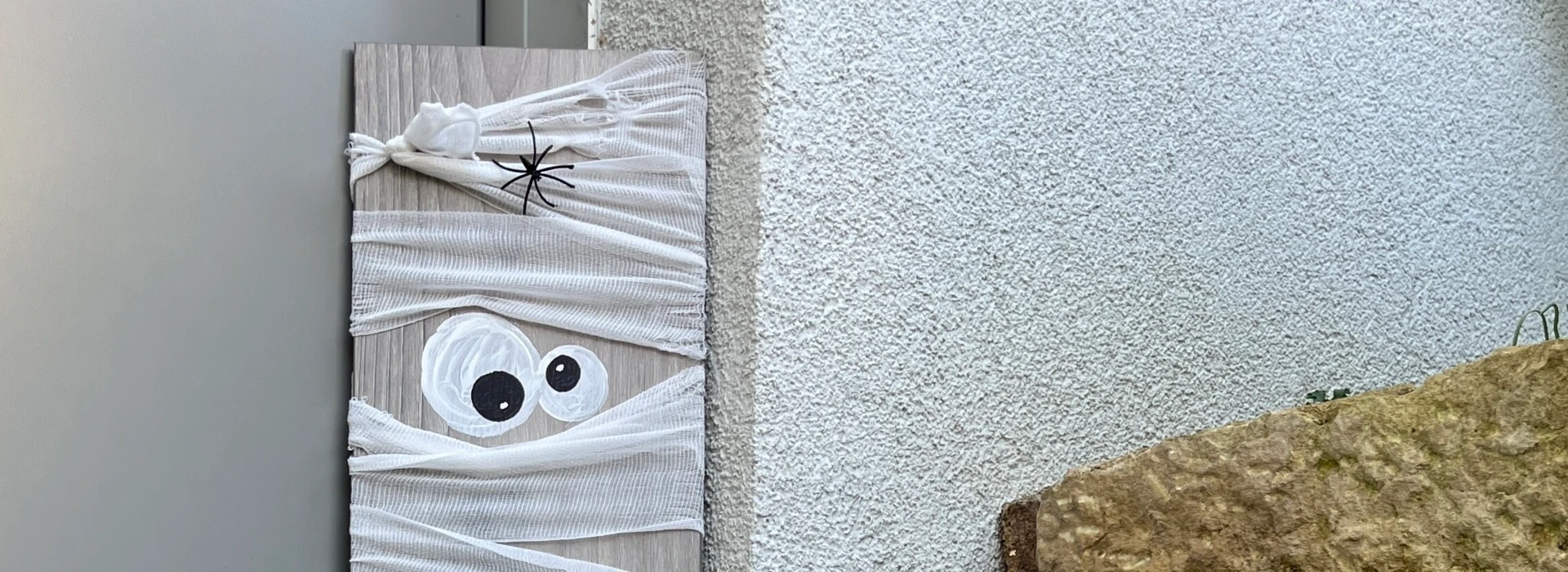 Eine DIY-Halloween-Dekoration mit Mumienfiguren aus Holz und Bändern, dekoriert mit Spinnen und Augen, neben einer Laterne im Freien.