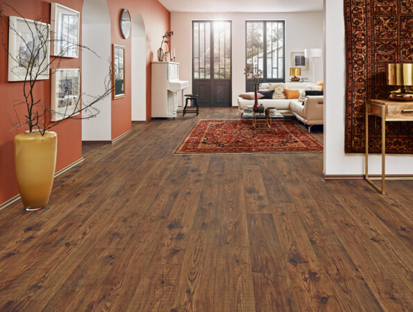 Spacious room with dark laminate flooring, featuring a piano, a sofa, and decorative elements like a large vase and a patterned rug.