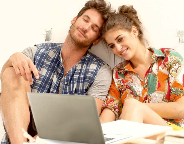 A couple sits together, smiling at a laptop screen, in a relaxed setting.