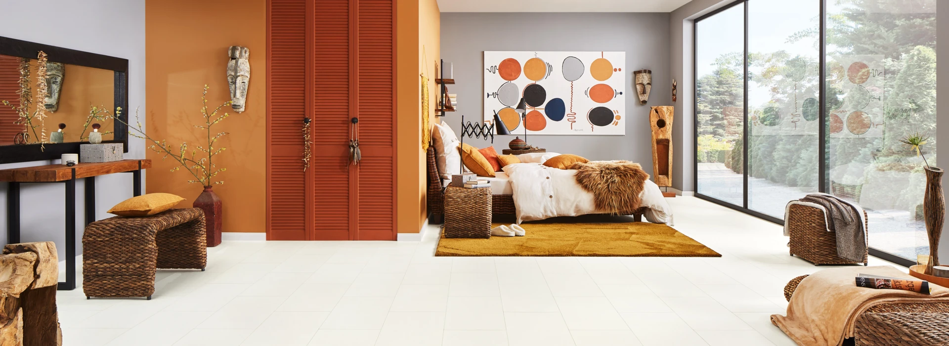 A bedroom with light laminate flooring, wicker furniture, and large windows, featuring orange accents and abstract wall art.