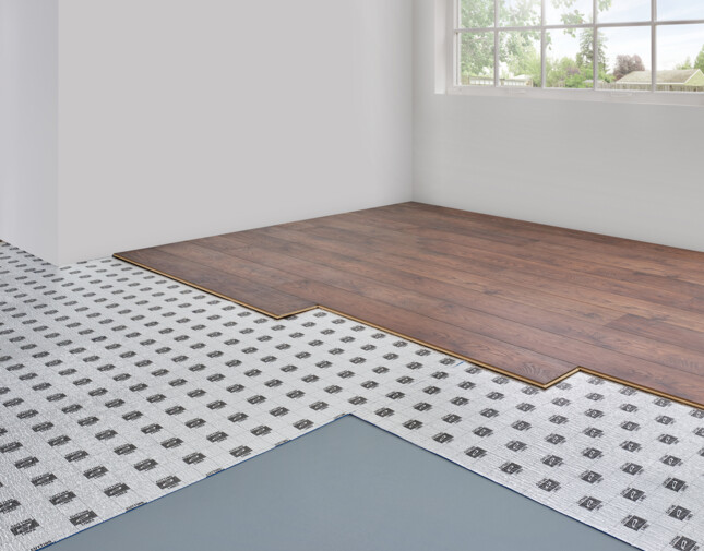 Laminate flooring installation showing layers, including underlay, in a bright room with a large window.