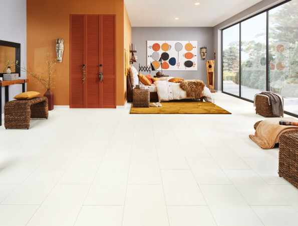 A bedroom with light laminate flooring, wicker furniture, and large windows, featuring orange accents and abstract wall art.