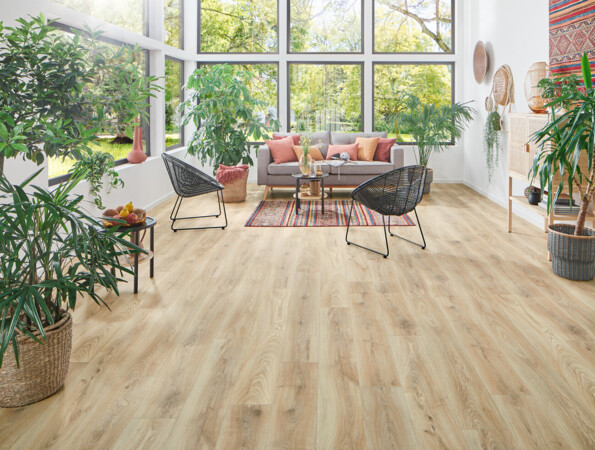 Wohnzimmer mit großen Fenstern, Pflanzen und einem Laminatboden. Gemütliche Einrichtung mit Teppich, Sofa und Stühlen.