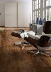 Helles Zimmer mit großem Fenster, braunem Laminatboden und gemütlichem Sessel. Im Hintergrund ist ein Bett mit Blick auf das Meer zu sehen.