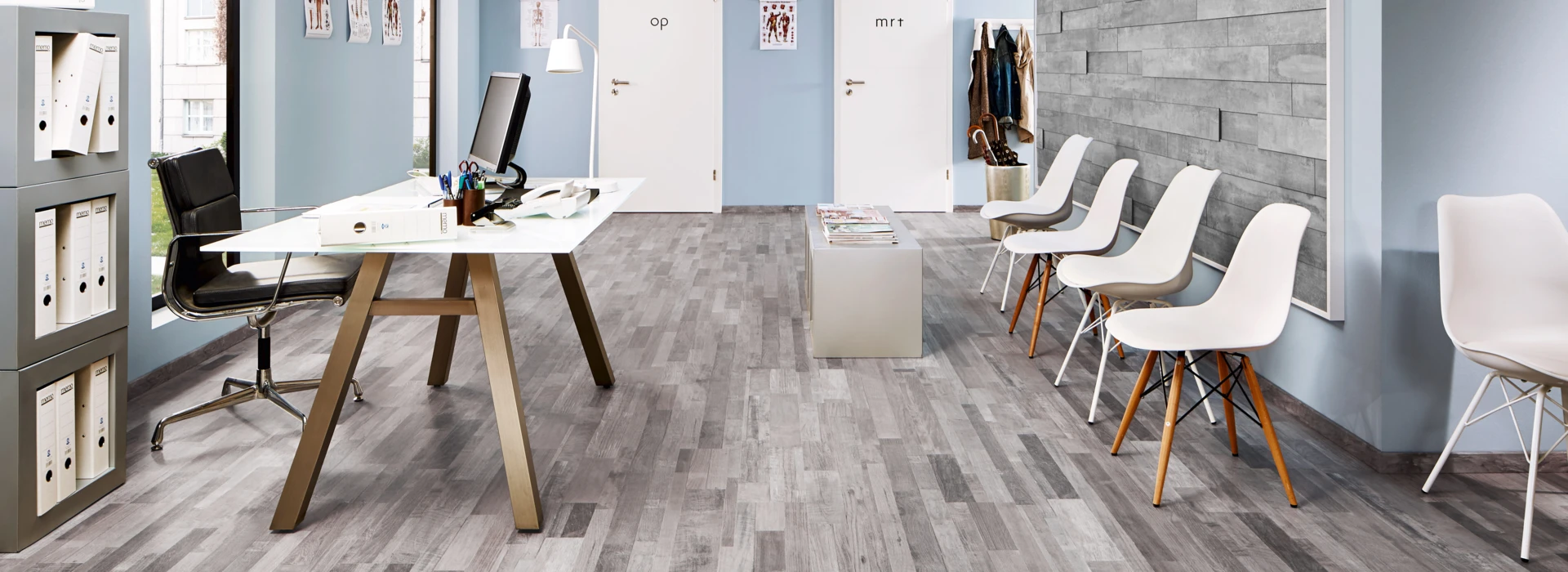 Office with grey laminate flooring, white desk, black chair, and waiting area with white chairs against blue walls.