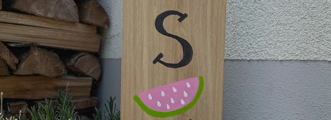 Holzschild im Garten mit der Aufschrift "hello SUMMER" und einer Wassermelonengrafik. Im Hintergrund sind Pflanzen und Holzstapel sichtbar.
