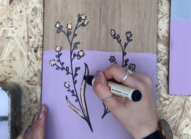 Eine Person zeichnet Blumenmuster auf ein Holzbrett, das teils lila bemalt ist.