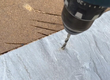 Eine Hand hält eine Bohrmaschine, die ein Loch in eine graue Platte bohrt.