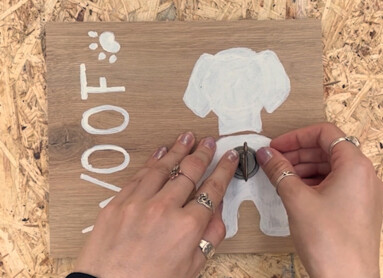 Person befestigt Haken auf Holzschild mit Hundemotiv und "WOOF" Schriftzug.
