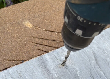 Eine Hand hält eine Bohrmaschine, die ein Loch in eine graue Platte bohrt.