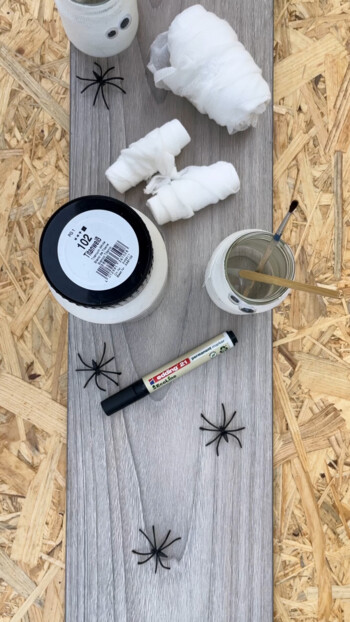 Bastelmaterialien auf Laminatboden, daneben Gläser, Spinnen, Marker und Mullbinden, für ein kreatives DIY-Projekt.