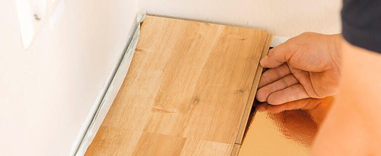 Ein Handwerker verlegt Laminatboden entlang einer Wand mit Steckdosen im Hintergrund.