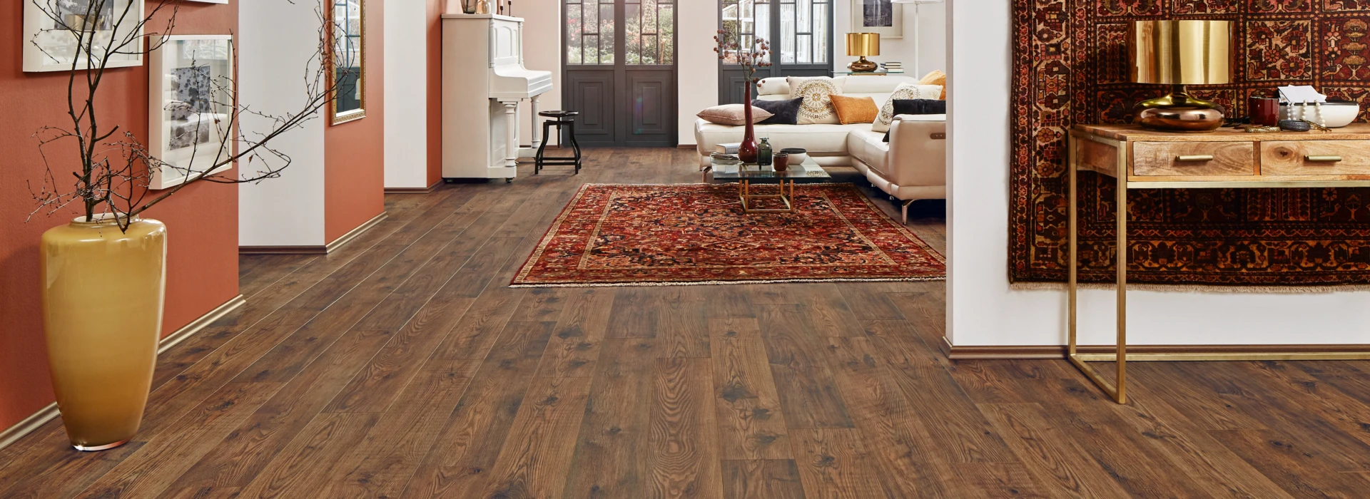 Spacious room with dark laminate flooring, featuring a piano, a sofa, and decorative elements like a large vase and a patterned rug.
