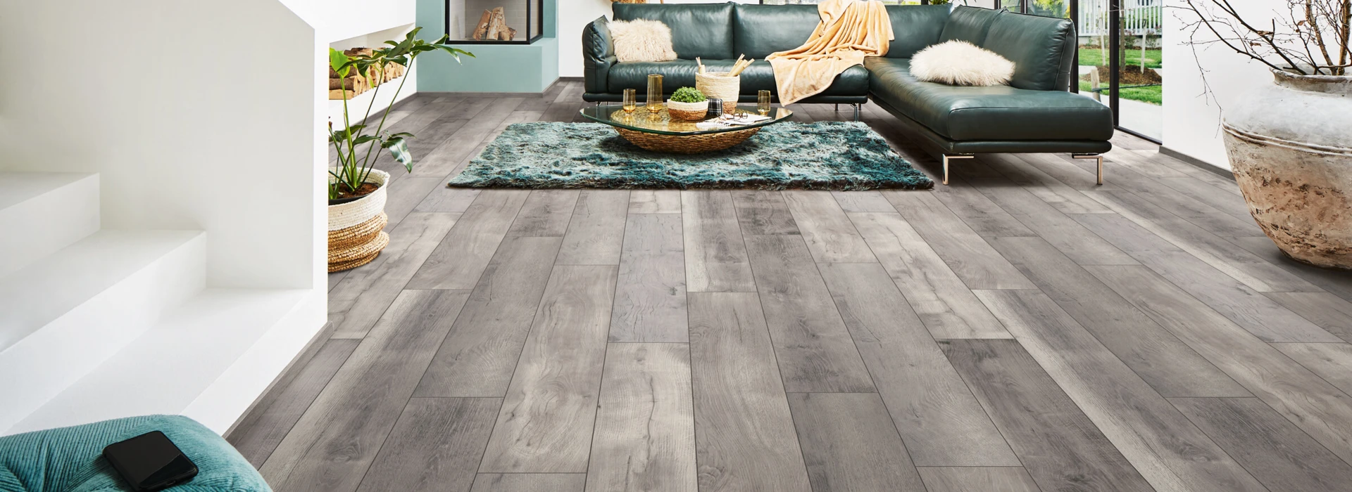 Modernes Wohnzimmer mit grauem Laminatboden der Serie 'Aquaprotect', grünem Sofa, großen Fenstern und stilvoller Dekoration.