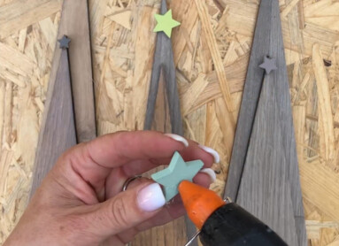 Eine Person dekoriert mit einer Heißklebepistole kleine Sterne auf holzfarbenen Dekorationselementen neben einer brennenden Kerze.