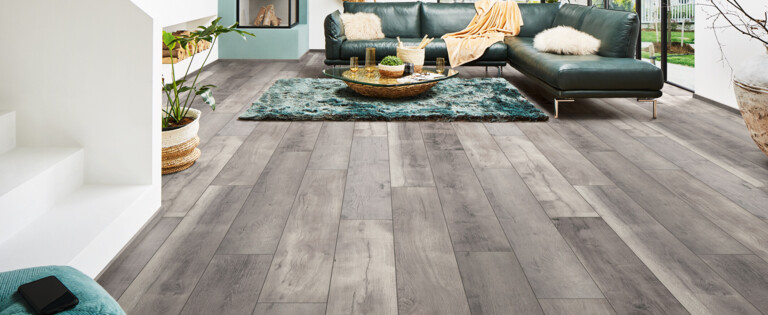 Modernes Wohnzimmer mit grauem Laminatboden der Serie 'Aquaprotect', grünem Sofa, großen Fenstern und stilvoller Dekoration.
