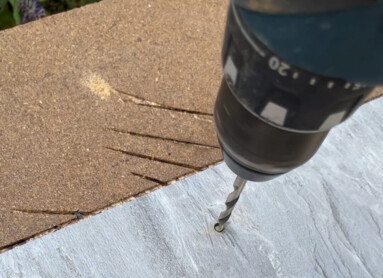 Eine Hand hält eine Bohrmaschine, die ein Loch in eine graue Platte bohrt.