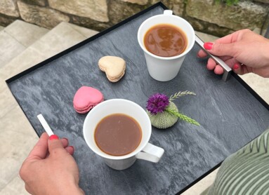 Eine Person trägt ein Tablett mit zwei Tassen Kaffee und Macarons im Freien, umgeben von grünen Pflanzen.