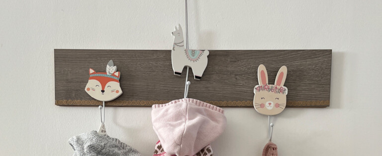 Kinderkleidung an Wandhaken über einer Holzkommode mit Kinderschuhen und grauem Fellkissen.