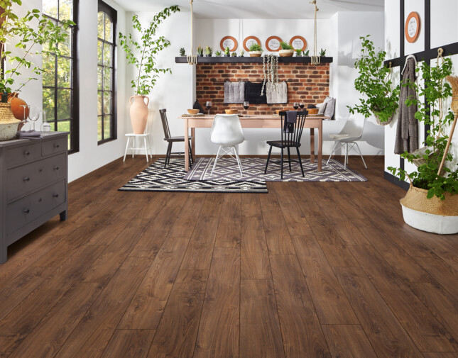 Spacious room with laminate flooring, white walls, large windows, plants, and a dining area featuring a brick accent wall and stylish decor.
