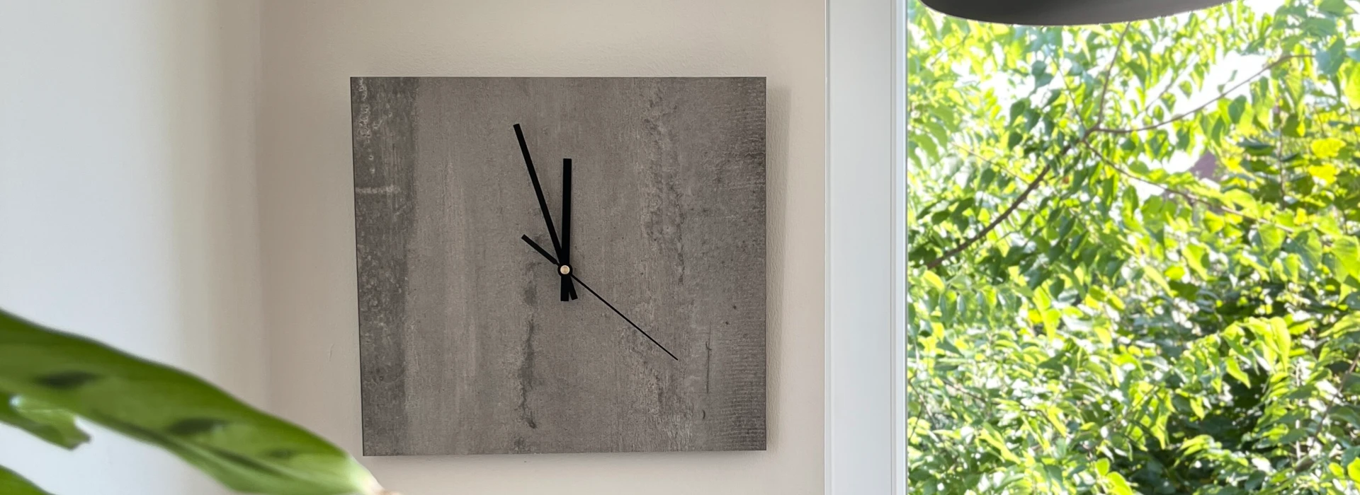 Gemütliche Ecke mit Kissen, Wanduhr und Pflanzen, neben einem Fenster mit Blick ins Grüne.