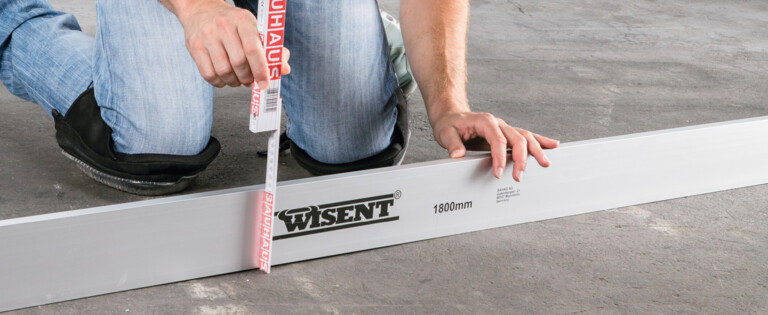 Ein Mann misst mit einem Maßband auf einem Betonboden, neben einem 1800mm langen Aluminiumprofil.