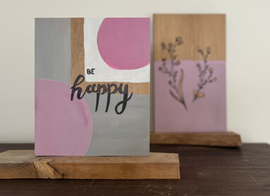 Zwei bemalte Holztafeln auf einem Tisch, eine mit der Aufschrift "Be Happy" in Rosa und Grau.