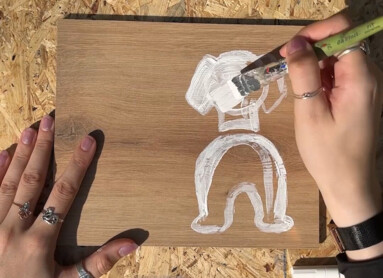 Eine Person bemalt eine Holzplatte mit weißer Farbe auf einem Tisch. Im Hintergrund steht ein großer Farbeimer.