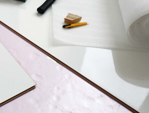 Tools and materials for installing laminate flooring, including a hammer, tape, and pencil, on a white surface.