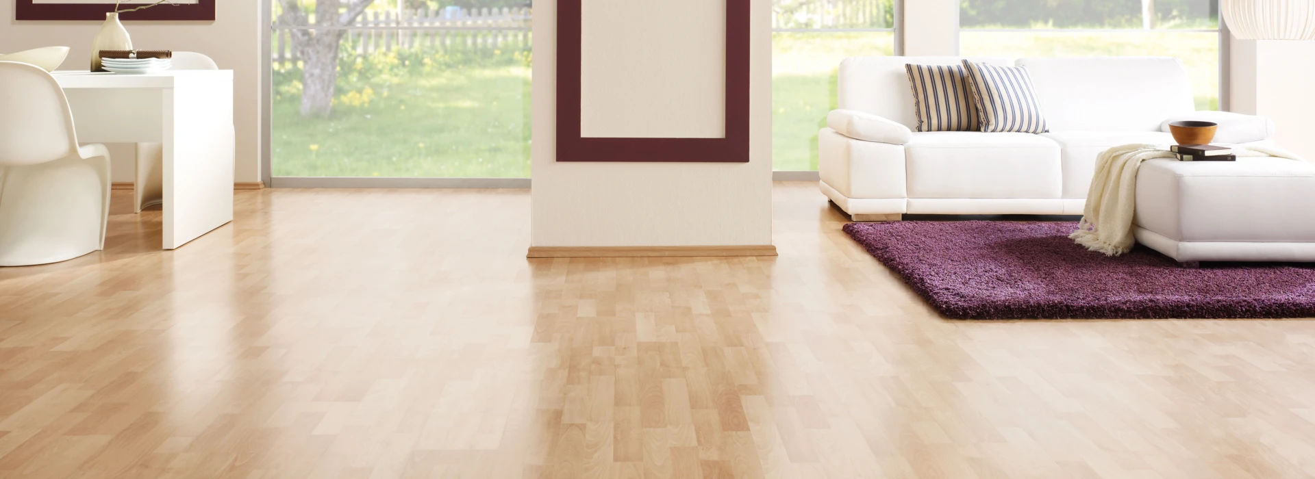 Living room with light laminate flooring, white furniture, and large windows overlooking a garden.