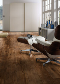 Helles Zimmer mit großem Fenster, braunem Laminatboden und gemütlichem Sessel. Im Hintergrund ist ein Bett mit Blick auf das Meer zu sehen.