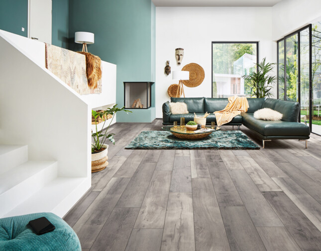 Modernes Wohnzimmer mit grauem Laminatboden der Serie 'Aquaprotect', grünem Sofa, großen Fenstern und stilvoller Dekoration.