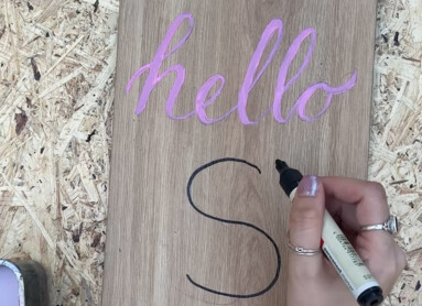 Eine Hand schreibt "S" auf ein Holzbrett mit "hello" in rosa Schrift.